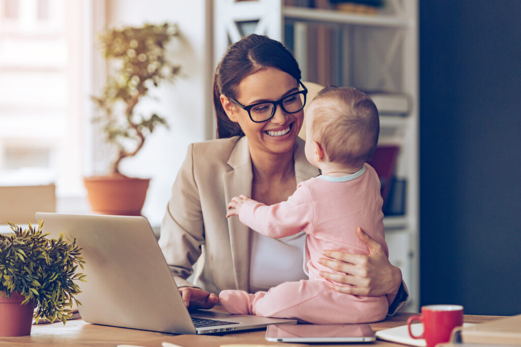 imprenditrice che lavora al laptop tenendo in braccio il figlio piccolo: esempio concreto di come un blocco del sito produttivo costringe a riorganizzare vita e lavoro familiare